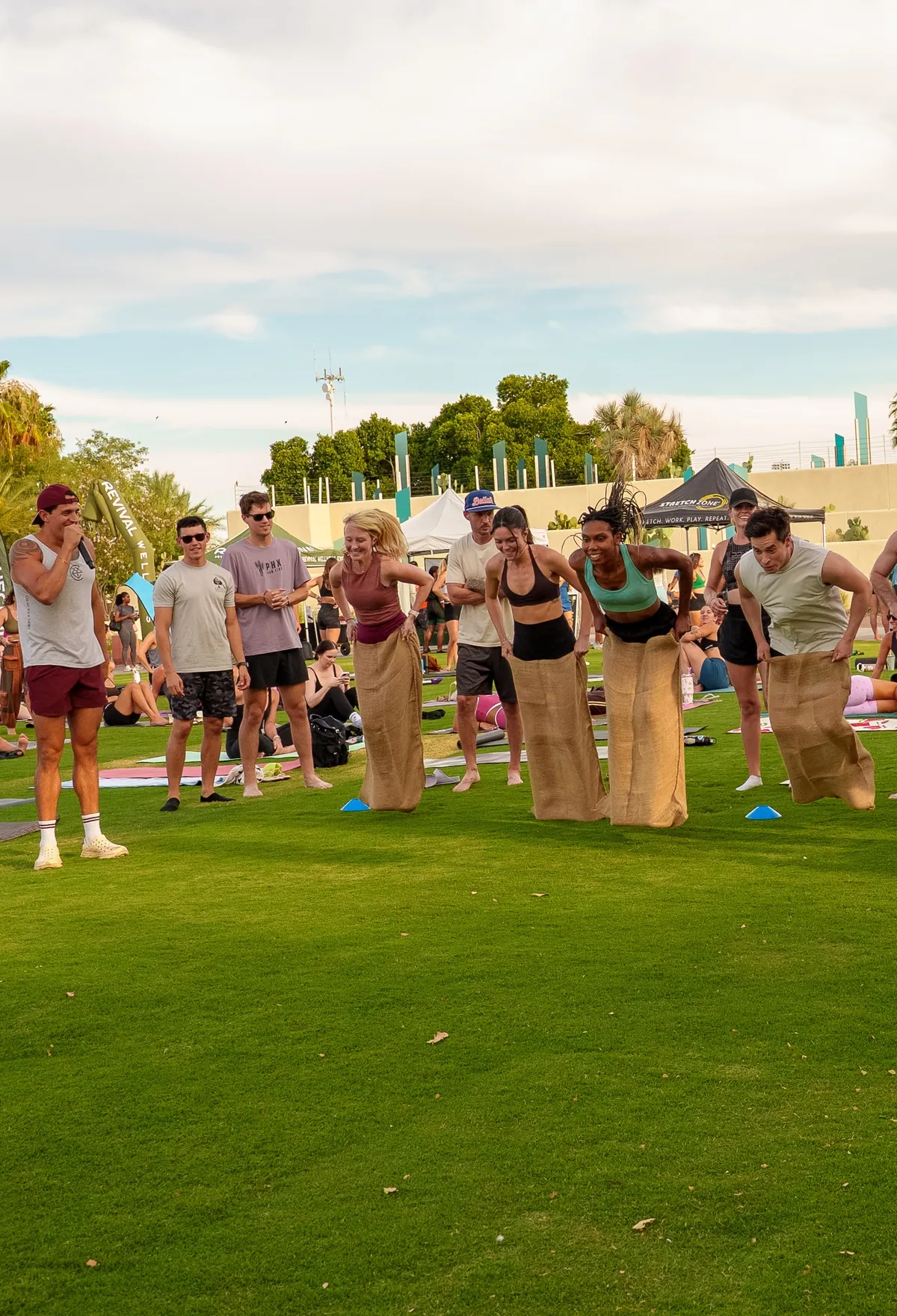 Four friends jumping in a sack race while others cheer from the lawn at a Revival field day