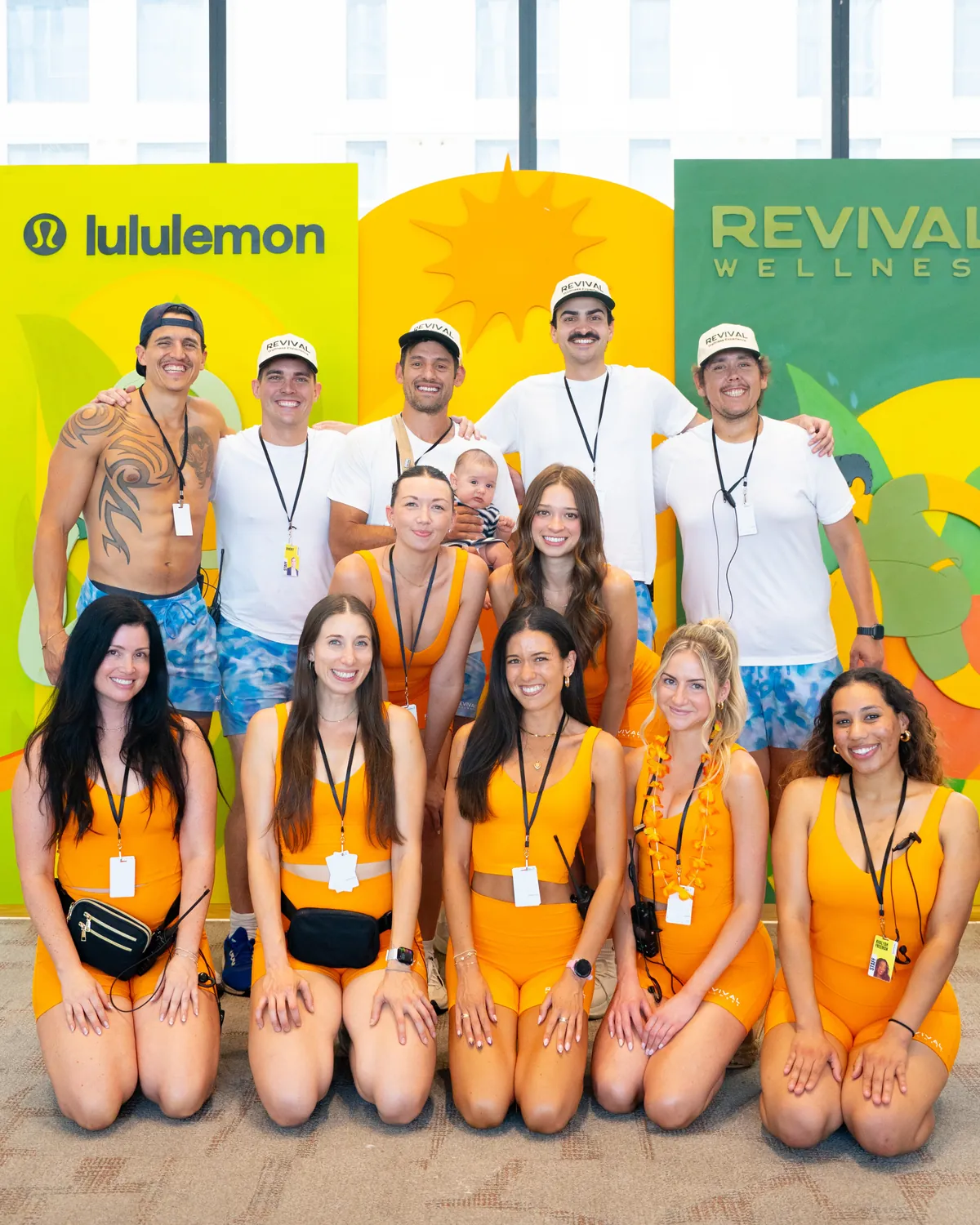 The Revival team in orange outfits and white shirts, with a baby, in front of a colorful Revival Wellness and lululemon backdrop